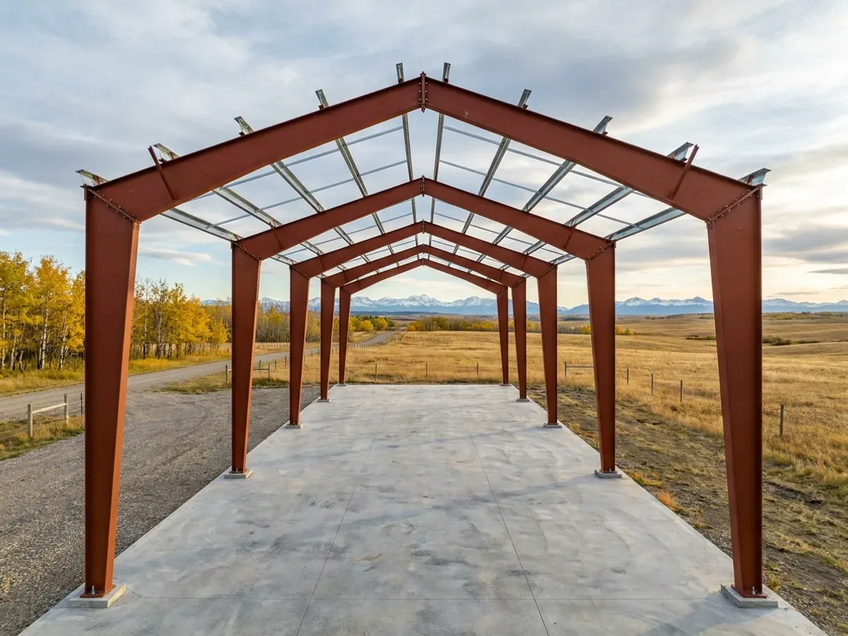 A red oxide primed structural steel frame partly erected on a concrete slab, showing bolted knee joints and anchor bolts, with autumn aspens behind.